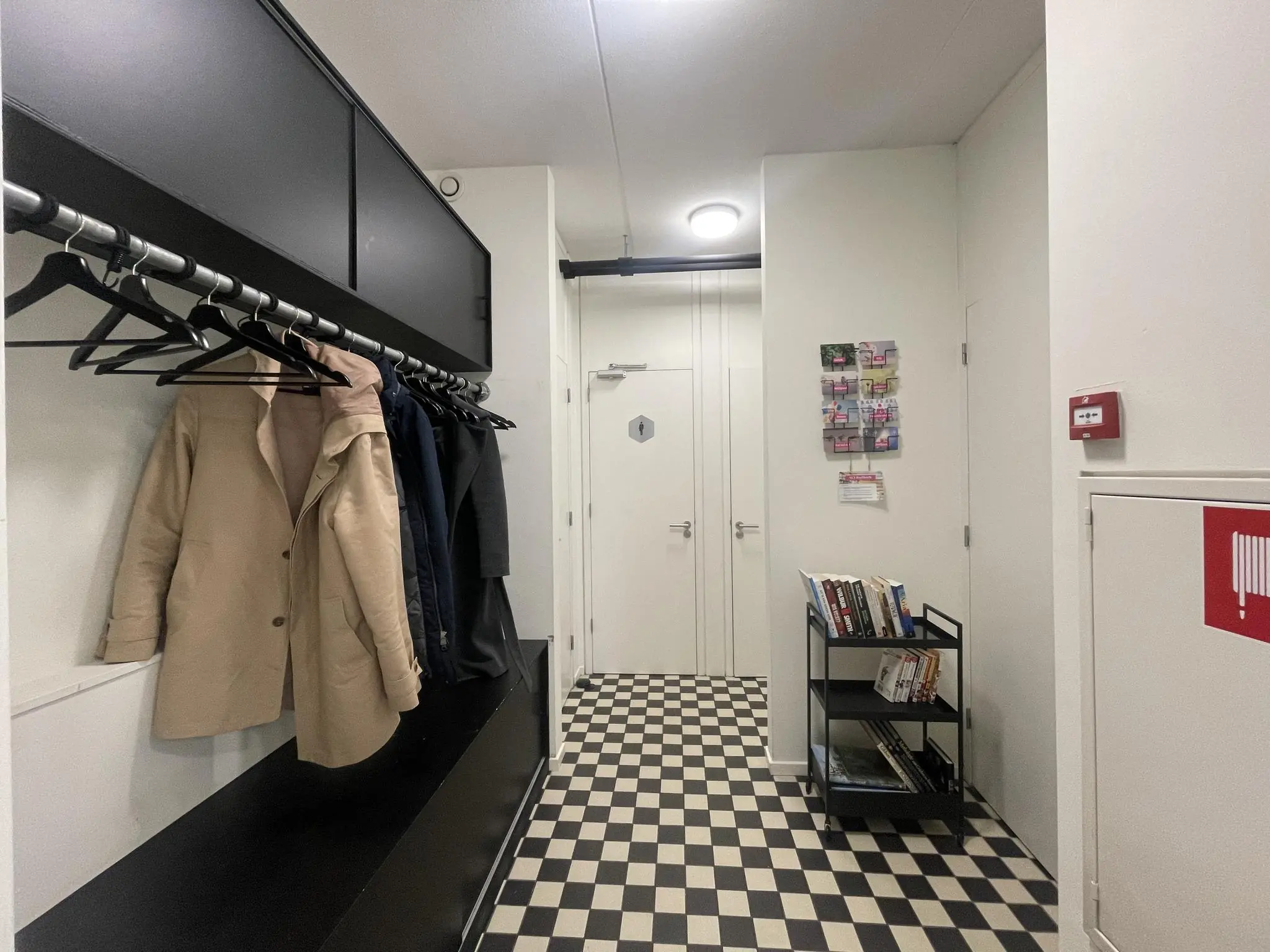 Hal met zwart-wit geblokte vloer, kapstok met jassen, boekenkar en folderrek aan de Jan Leentvaarlaan.